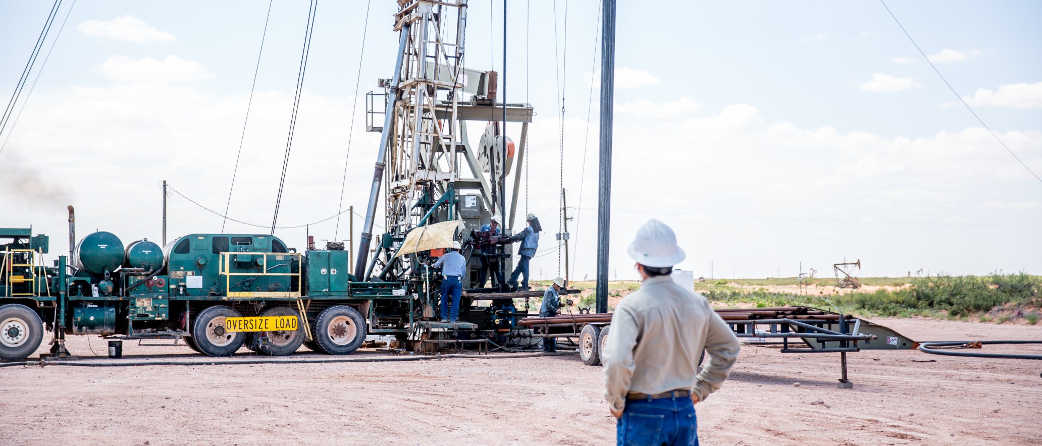 A worker, wearing a white hard hat and protective gear, stands at a distance observing a drilling rig operation at an industrial site. The machinery, vehicles, and equipment are set up on a dirt lot with open sky and distant structures in the background.