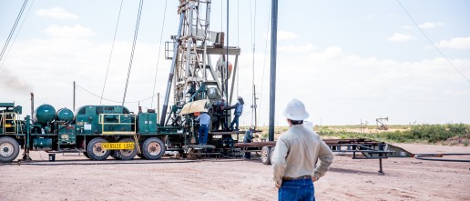 A worker, wearing a white hard hat and protective gear, stands at a distance observing a drilling rig operation at an industrial site. The machinery, vehicles, and equipment are set up on a dirt lot with open sky and distant structures in the background.